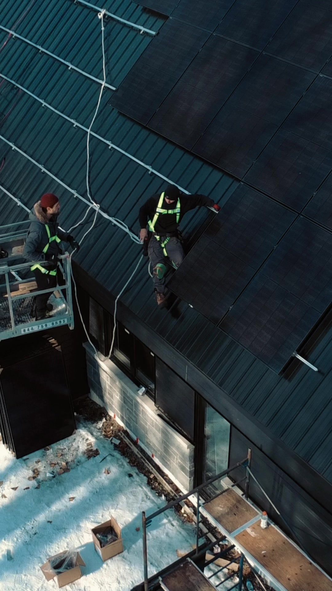 Deux installateurs de Larks Solar en harnais, l'un sur un toit d'acier couvert de panneaux, l'autre dans une nacelle, en hiver à Saint-Calixte
