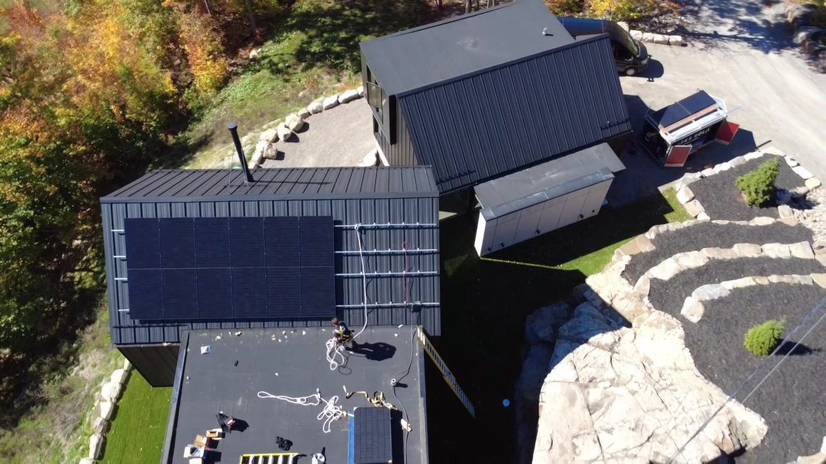 Vue de drone d'une maison à Sainte-Adèle : panneaux noirs sur le toit d'acier, installateur sur le toit plat voisin, forêt d'automne autour